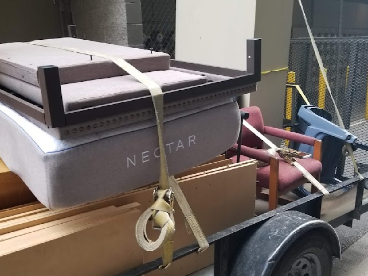 A trailer loaded with furniture, including a mattress, chair, and boxes, secured with straps.