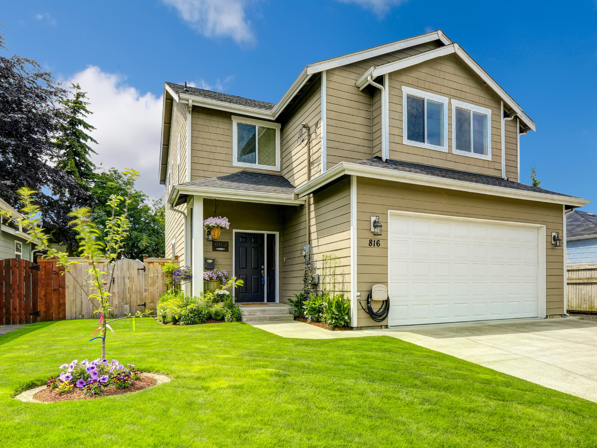 Two-story house with tan siding, white garage door, and green lawn under a blue sky.