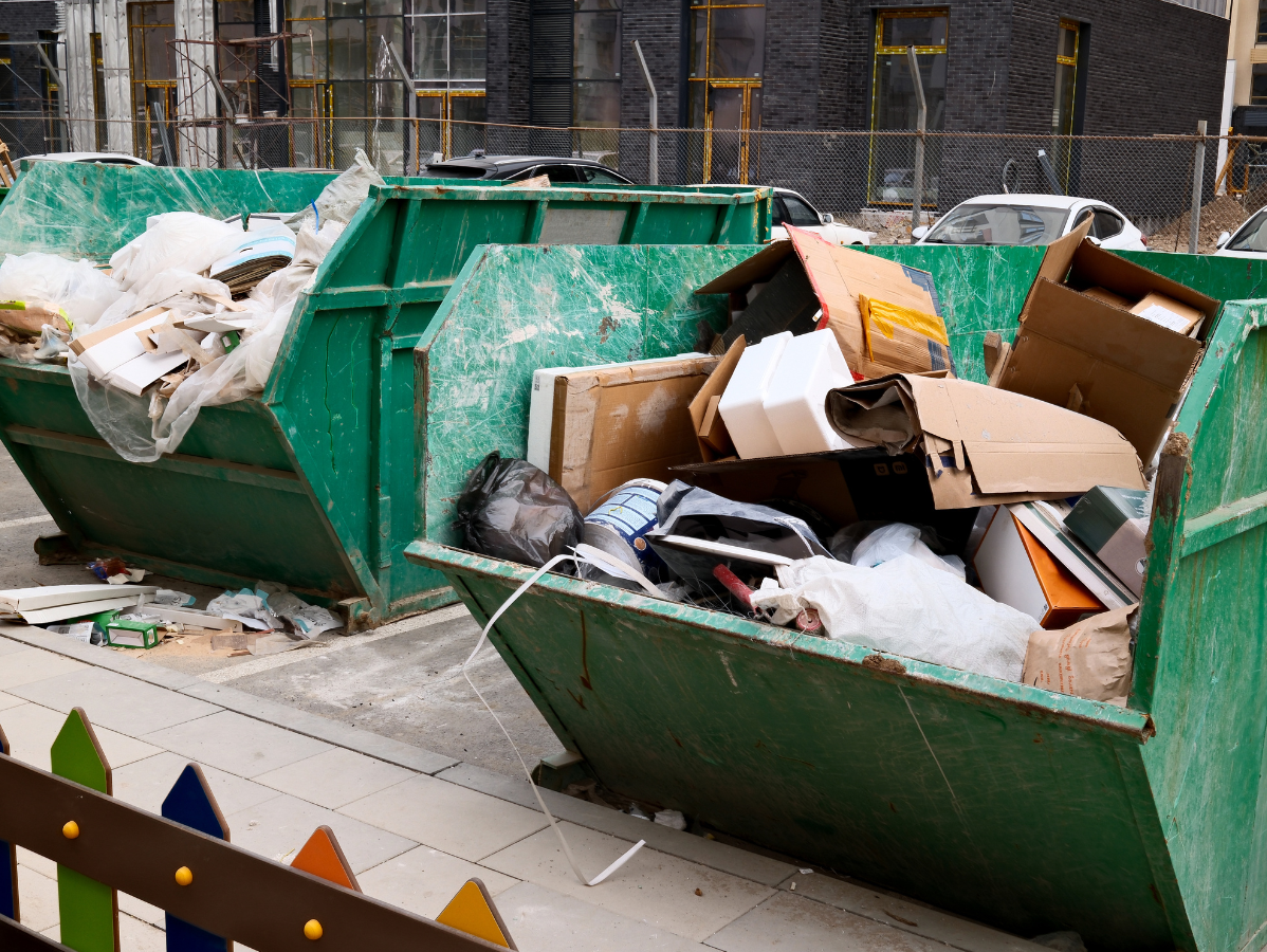 Two overflowing green dumpsters on a paved area, containing trash and debris.
