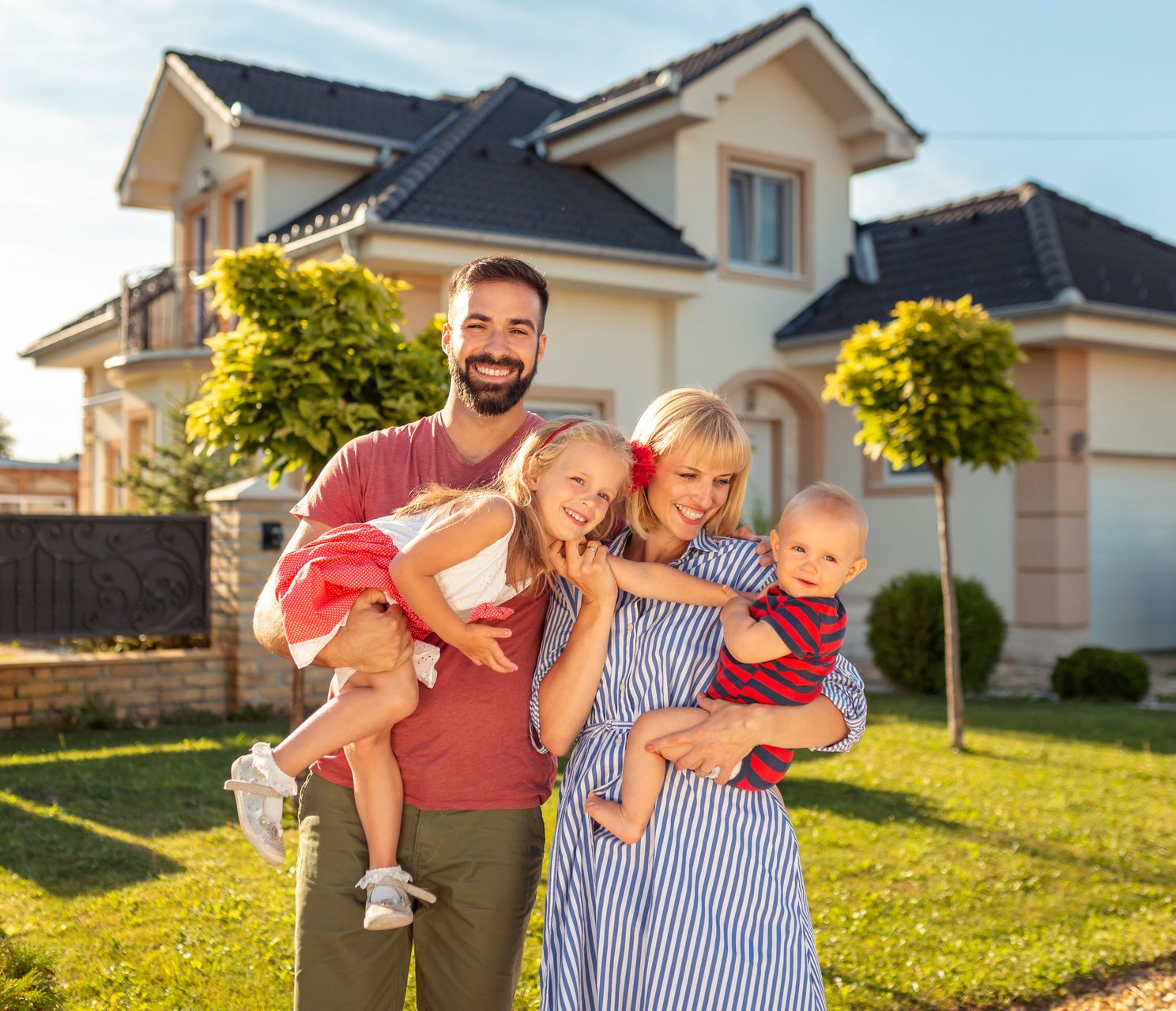 Family smiling in front of a house, with parents holding three young children on a sunny lawn.