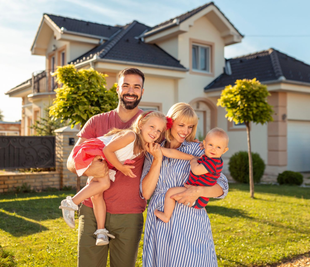 Family smiling in front of a house, with parents holding three young children on a sunny lawn.