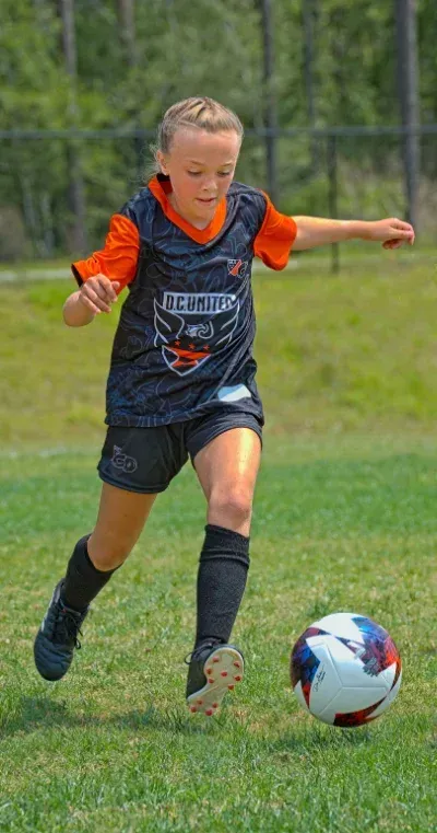 A young girl is kicking a soccer ball on a field dressed in MLS Go Coppermine  Recreation League DC United uniform.