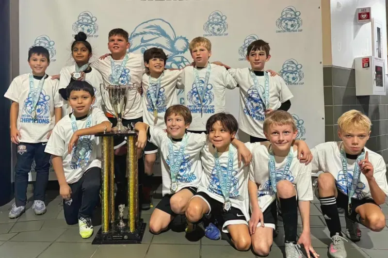 Youth soccer team posing with a trophy in a locker room, wearing white and blue uniforms.