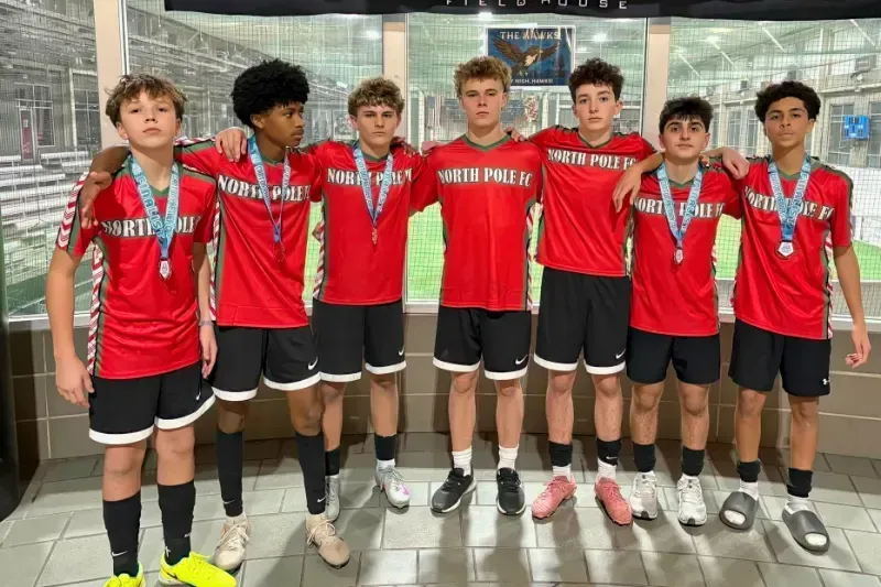 Seven boys in red jerseys and medals pose together in front of a stadium backdrop.