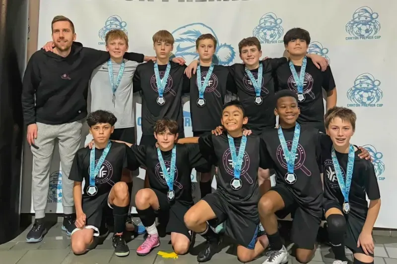 Youth soccer team posing with medals in front of a backdrop, wearing black uniforms and smiling.