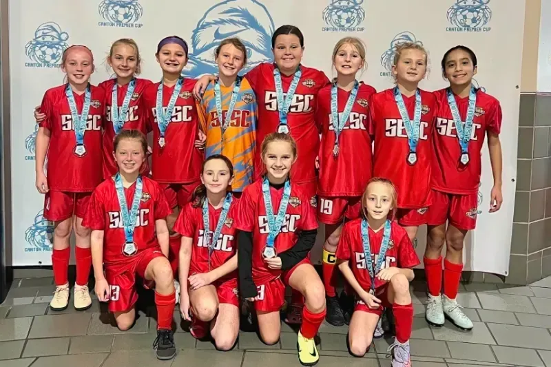 Youth soccer team in red uniforms posing with medals in front of a sponsor backdrop