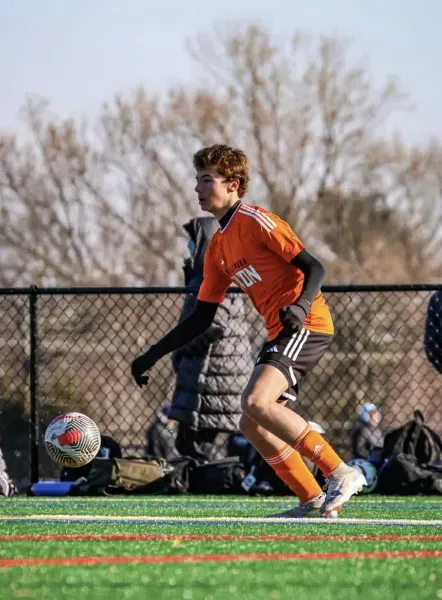 Soccer player in orange jersey dribbles on a field with a chain-link fence and trees in the background