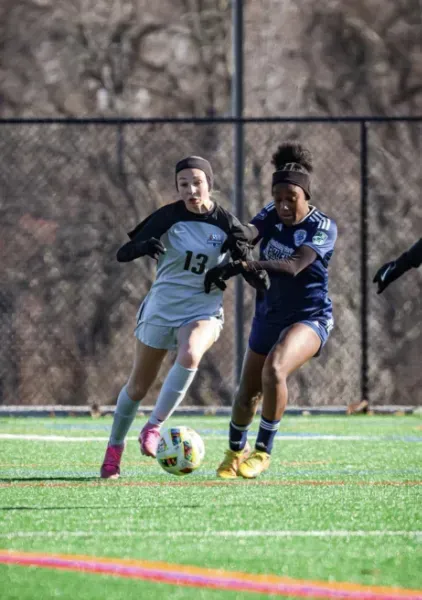 Two soccer players in white and blue jerseys chase the ball on a sunny field