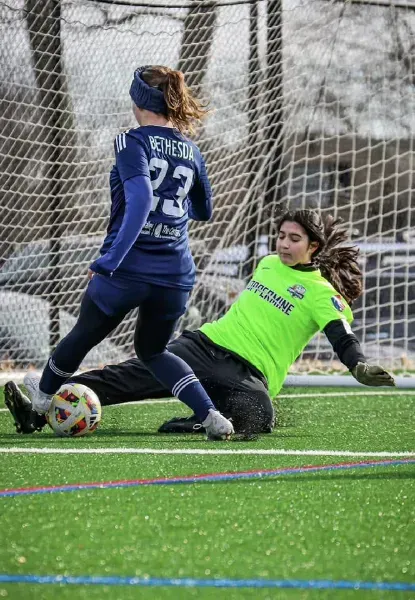 Soccer player in blue challenges goalkeeper in neon green near the goal on turf