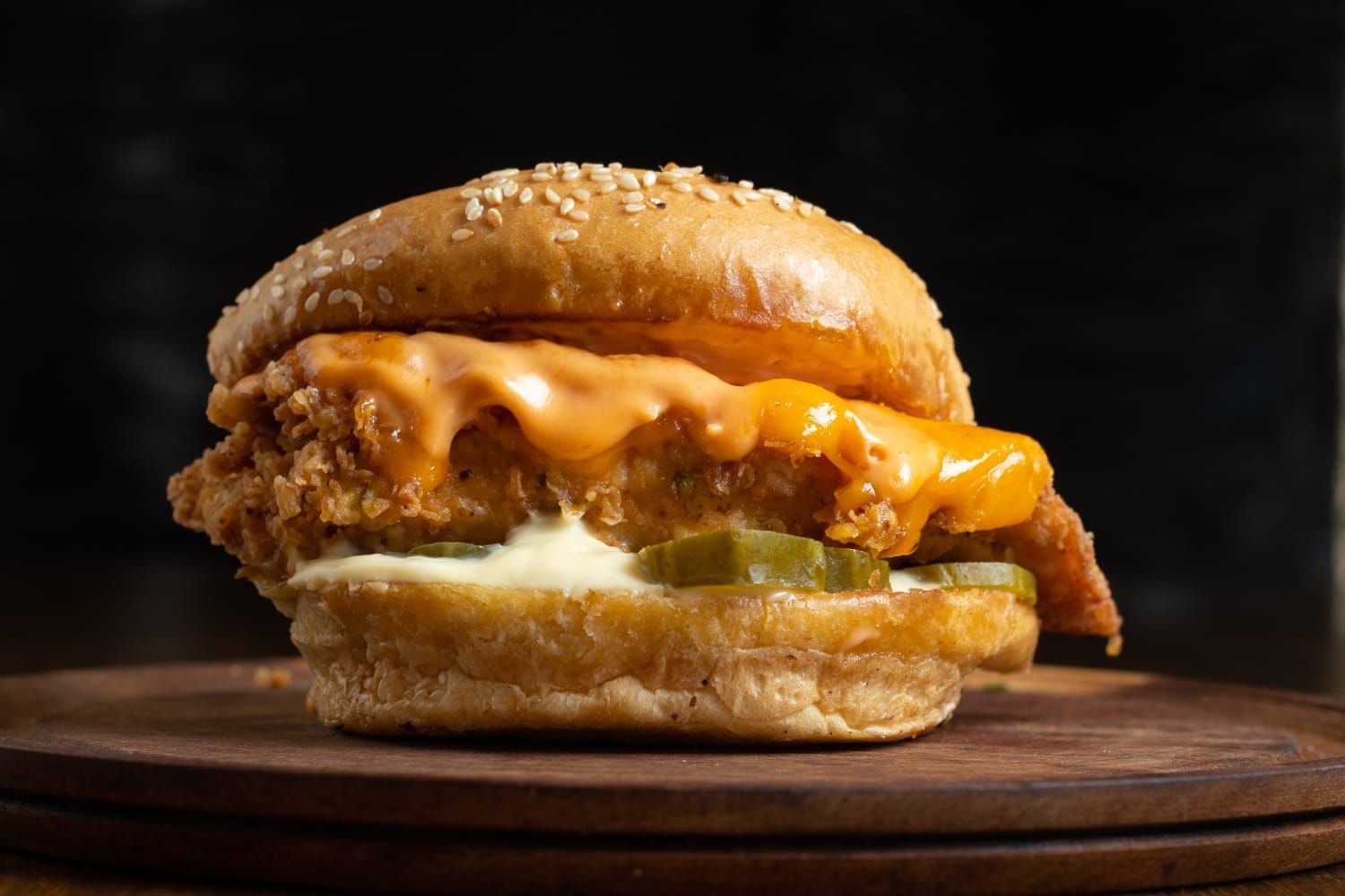 Sándwich de pollo frito sobre una tabla de madera, cubierto con queso y pepinillos.