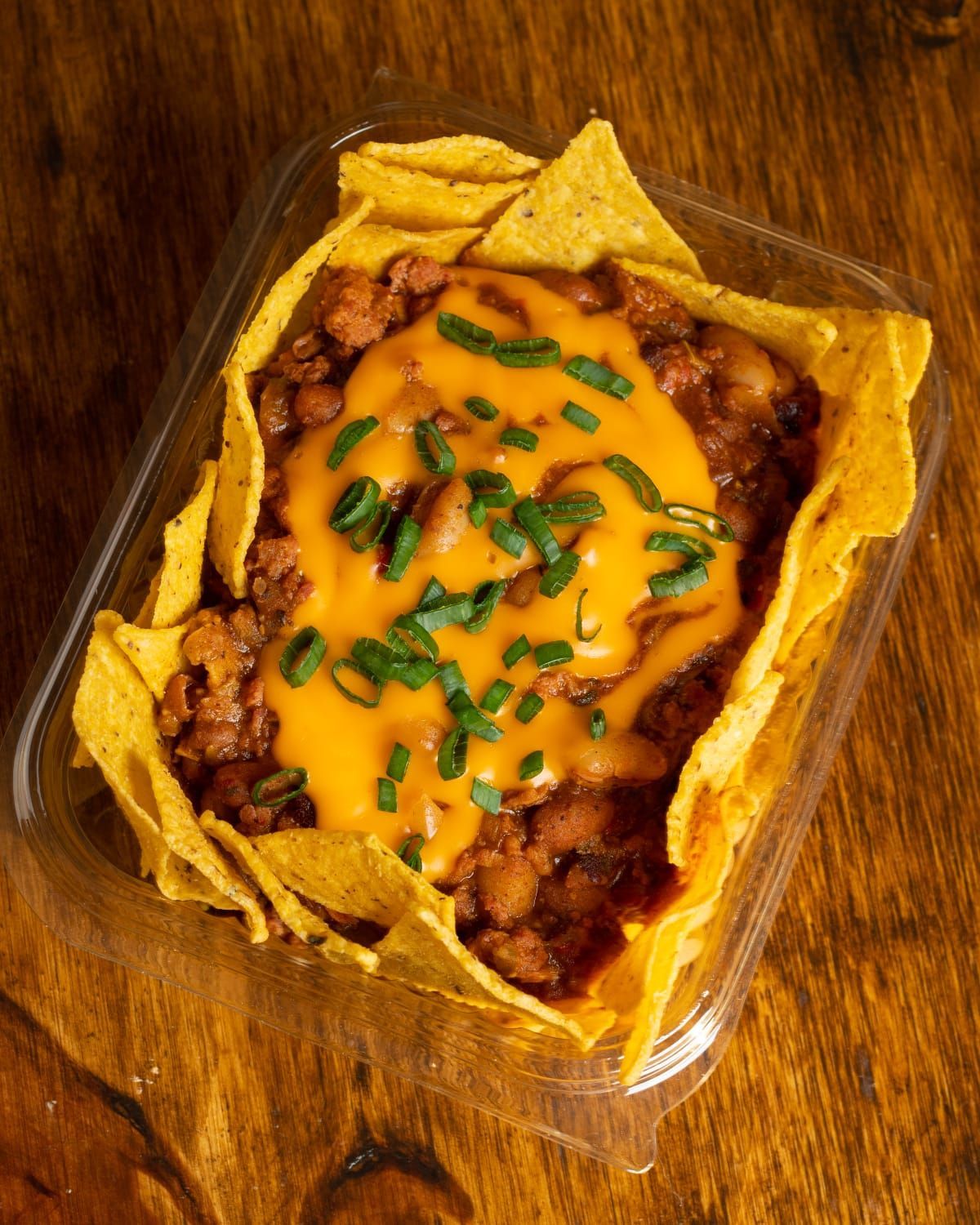 Nachos con chile y queso en un recipiente transparente, adornados con cebollino sobre una superficie de madera.