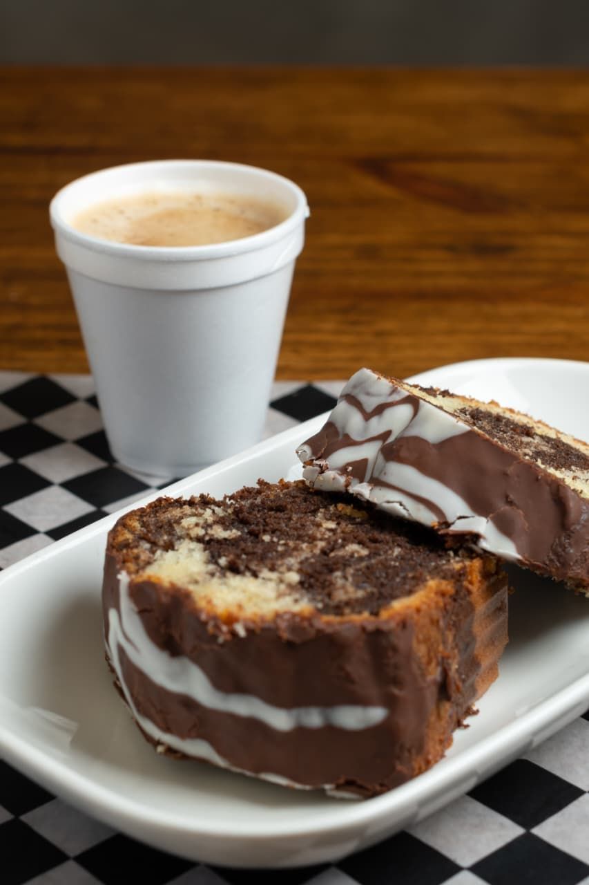 Rebanadas de pastel de chocolate marmolado con glaseado blanco y café en una taza blanca sobre una servilleta a cuadros.