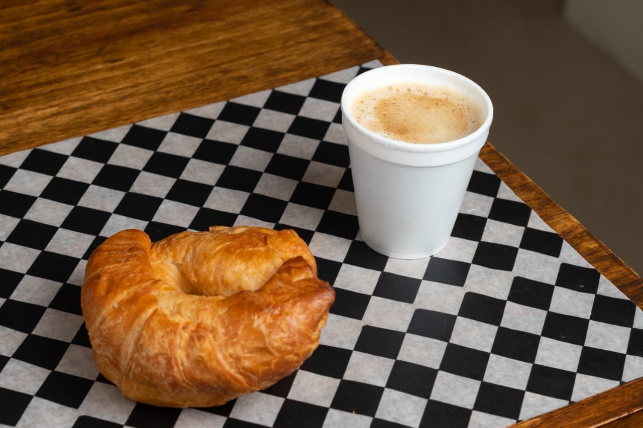 Croissant y café en una taza blanca sobre un papel a cuadros en blanco y negro sobre una mesa de madera.