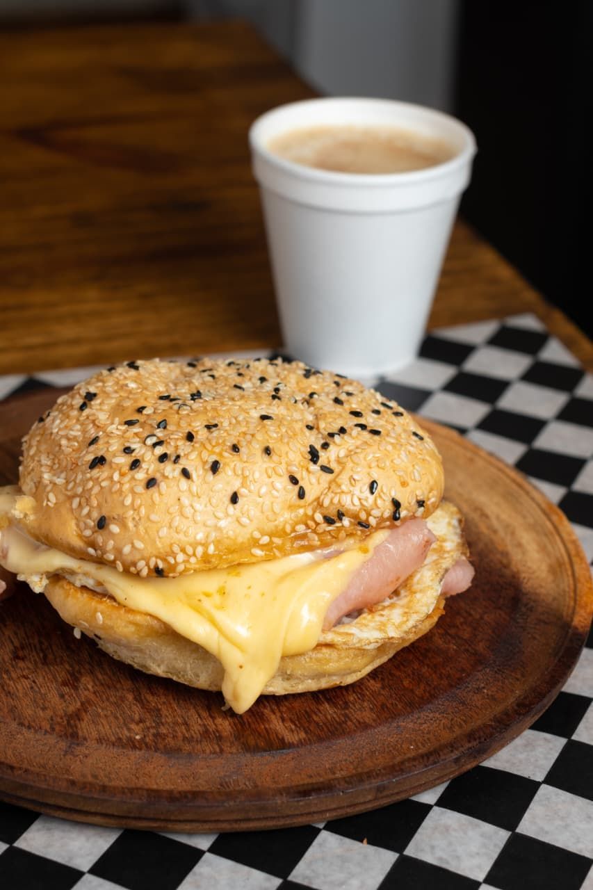 Un sándwich de bagel con semillas de sésamo, queso y jamón en un plato de madera, junto a una taza de café.