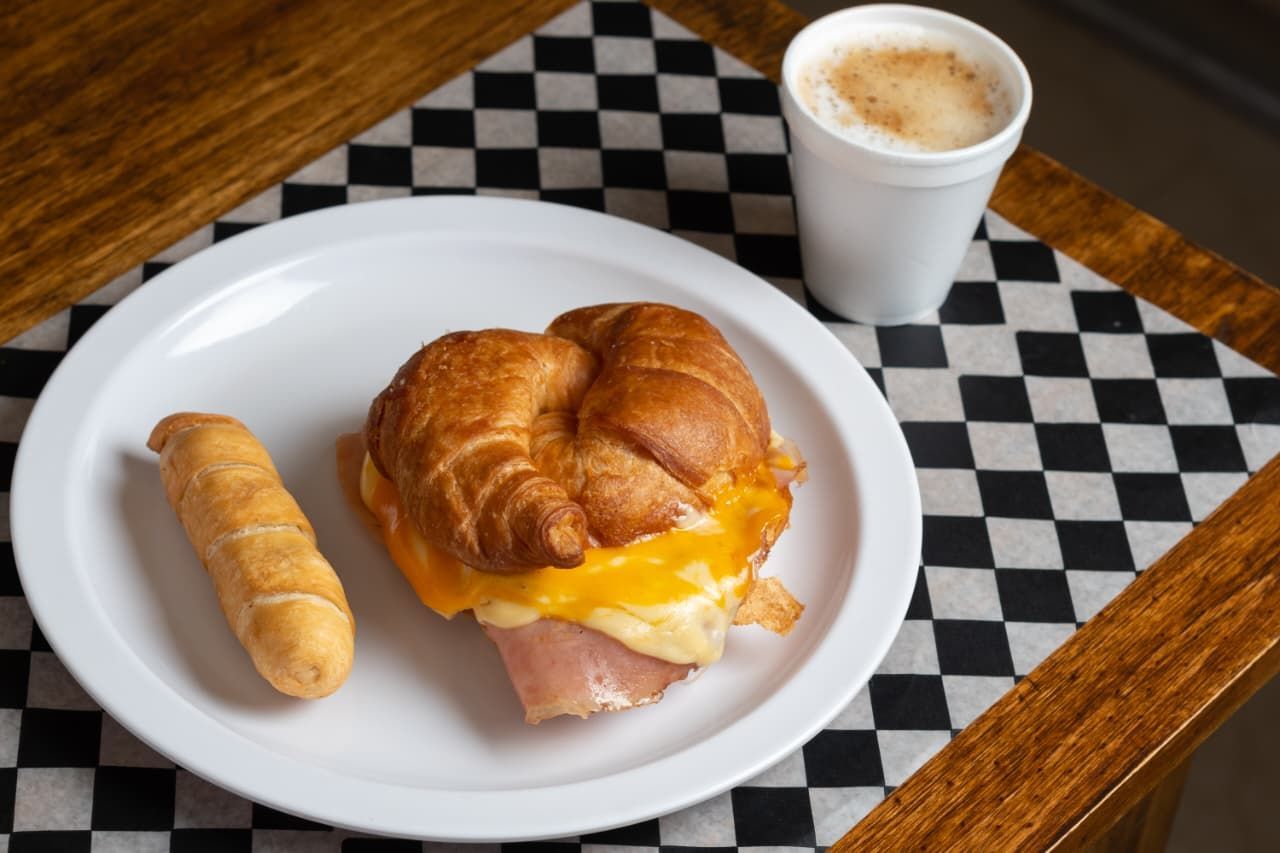 Desayuno en plato blanco con sándwich de croissant, pastelería y café.