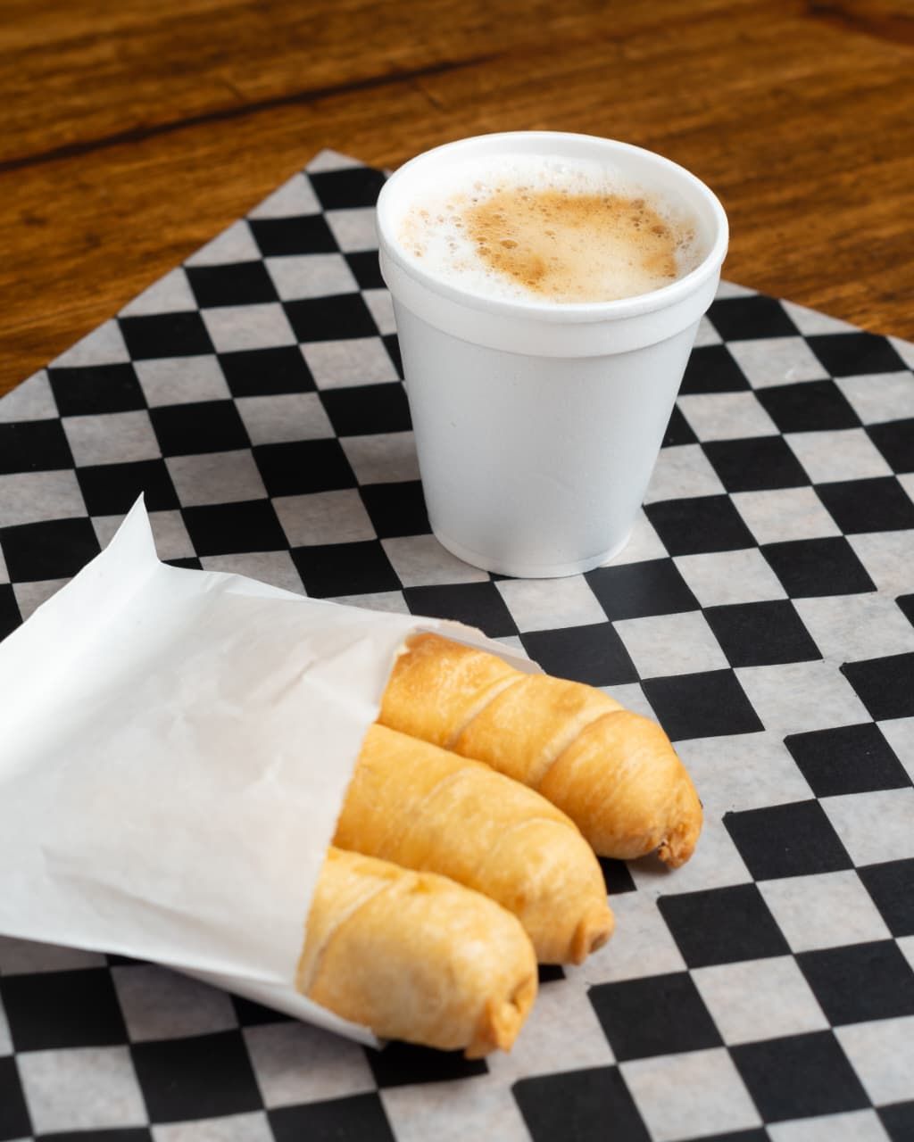 Tres pasteles fritos envueltos en papel con una bebida de café sobre una superficie a cuadros.