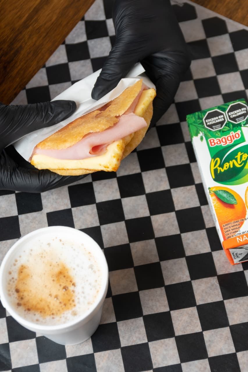 Persona sosteniendo un sándwich de jamón y queso junto a un café y jugo de naranja sobre una superficie a cuadros.