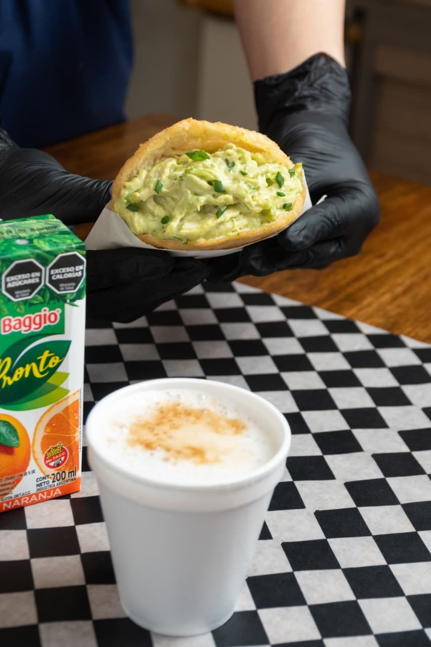 Persona con guantes negros sosteniendo una arepa rellena, una taza de capuchino y una caja de jugo de naranja
