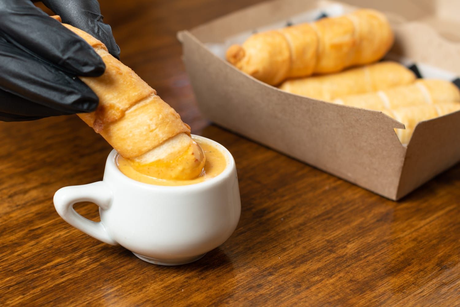 Una persona que lleva un guante negro sumerge un palito de queso frito en una taza de salsa de naranja