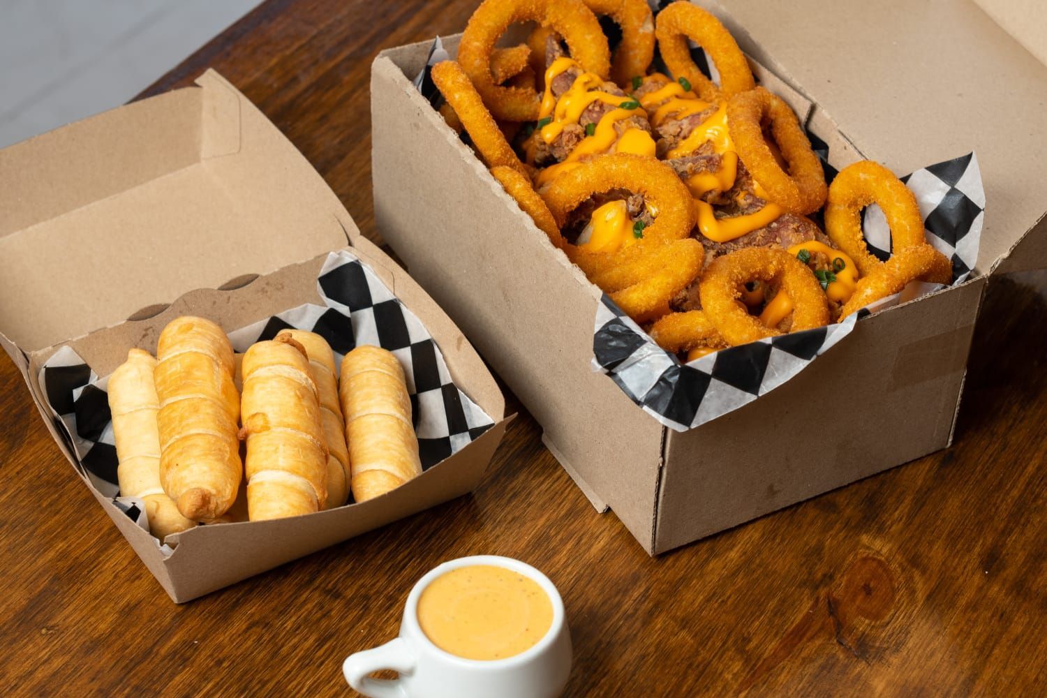 Cajas de comida frita con salsa para mojar: aros de cebolla, tequeños y salsa de queso sobre una mesa de madera.