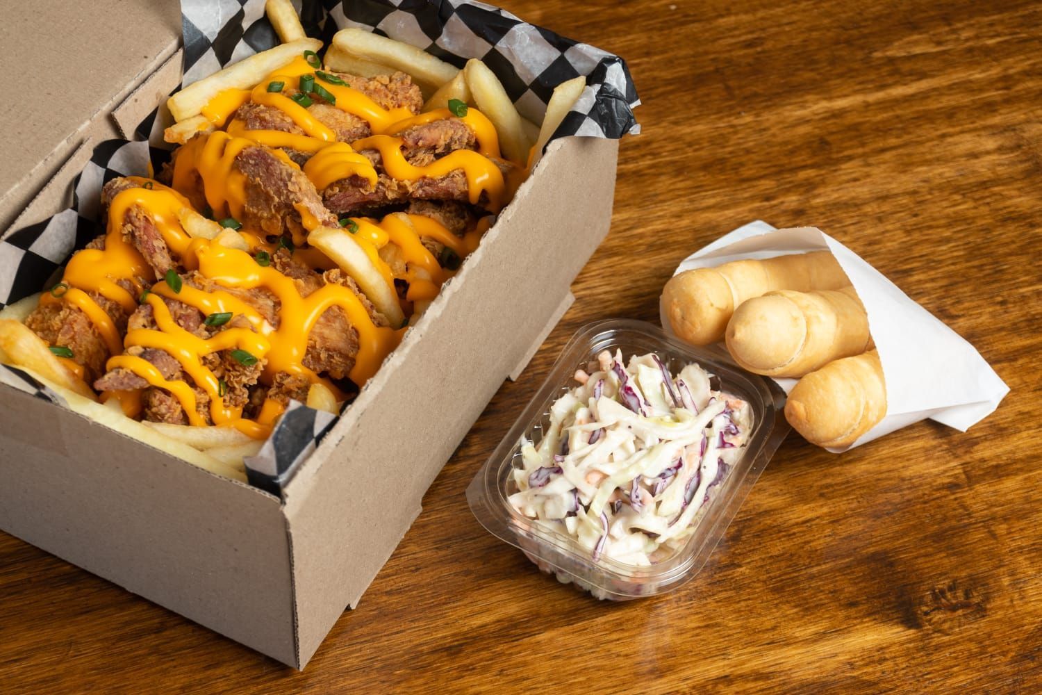 Caja de pollo frito y papas fritas con salsa de queso, ensalada de col y palitos de queso sobre una superficie de madera.