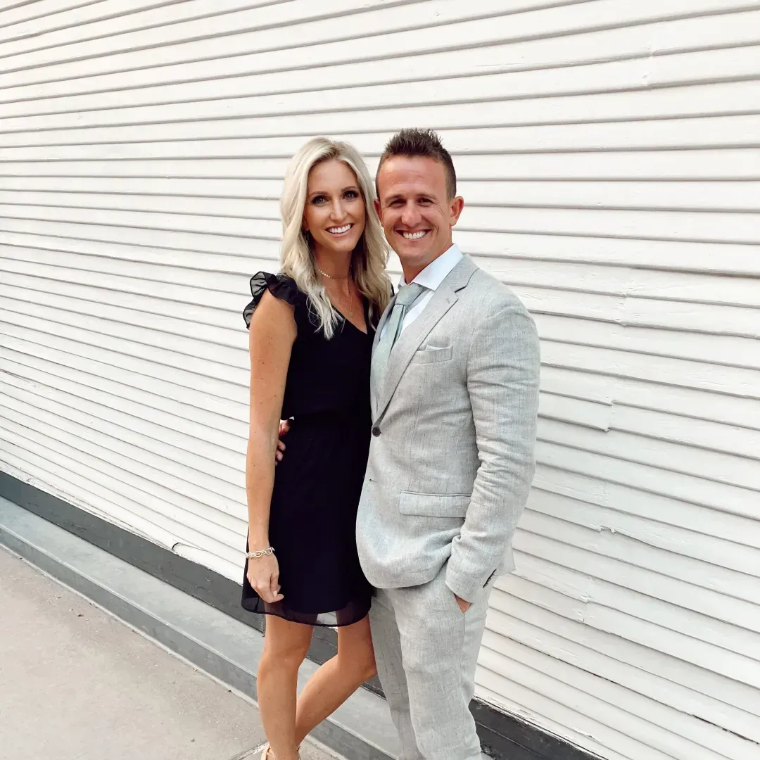 A man in a suit and a woman in a black dress are posing for a picture in front of a white wall.
