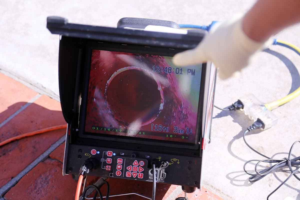 Monitor displaying a close-up view inside a pipe. A gloved hand points at the screen. Outdoors on concrete.