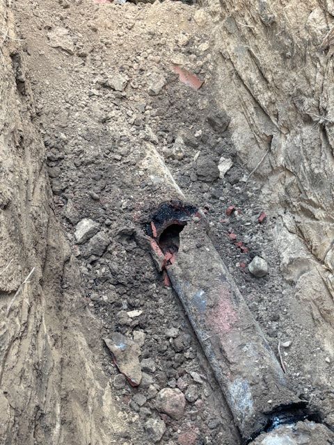 Broken pipe in an excavated trench, surrounded by dirt and brick fragments.