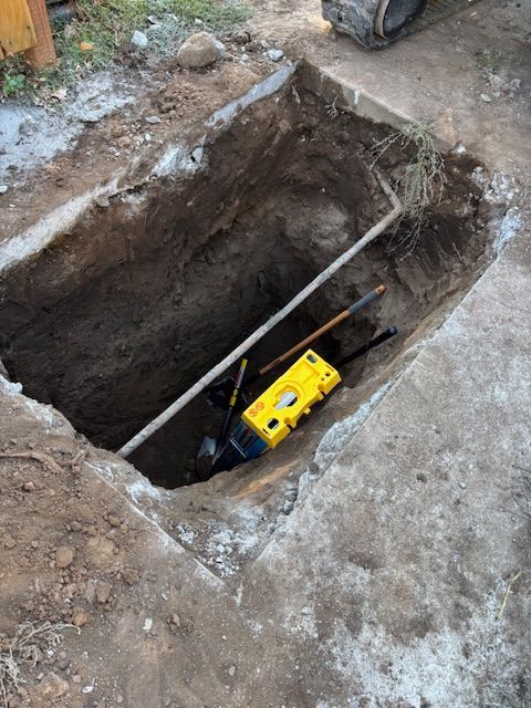 Trench dug in concrete, revealing pipe. Yellow device and tools inside.