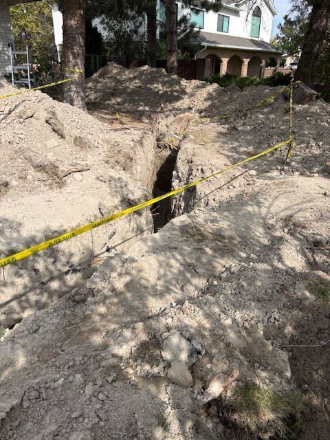 A deep trench dug in dirt, yellow caution tape strung around the perimeter; a house in the background.