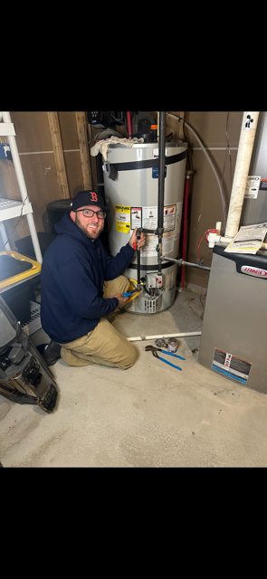 A person kneels near a water heater, pointing with a tool. They wear a blue hoodie and a baseball cap.