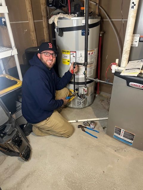 Man kneels near a water heater, holding pliers, smiling. Basement setting with a furnace visible.