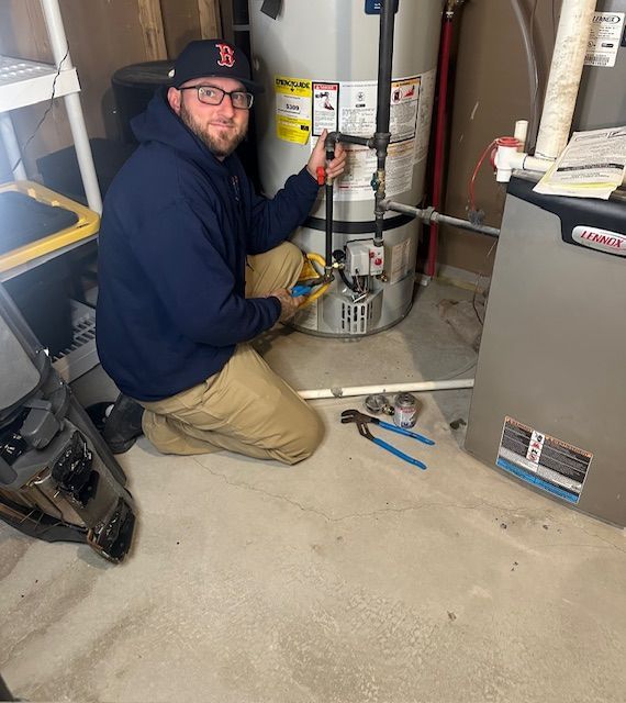 A person in a baseball cap works on a water heater in a basement, holding tools.
