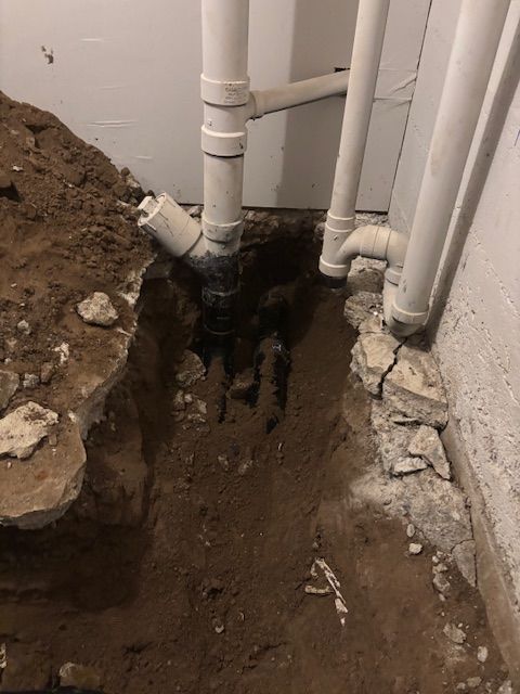 Trench dug near foundation exposing black plumbing pipes beneath white PVC pipes. Soil and debris are present.