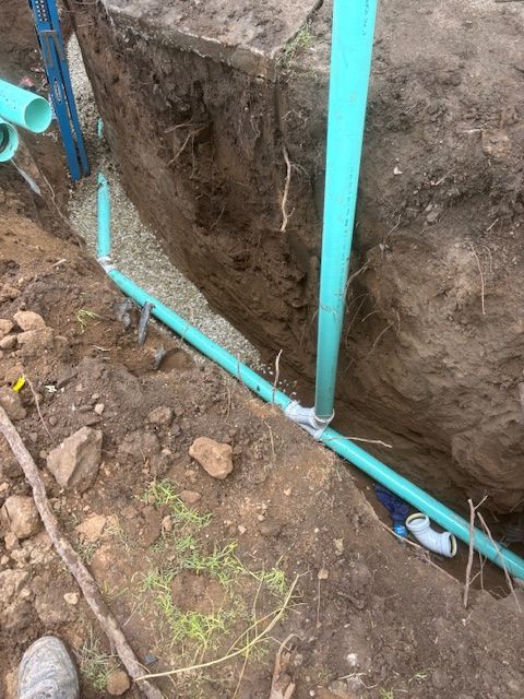 Blue PVC pipes in trench, connecting to a white fitting. Gravel and dirt surround the pipes.