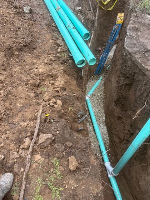 Trench with blue pipes and fittings. Some pipes are loose. Soil and gravel visible.
