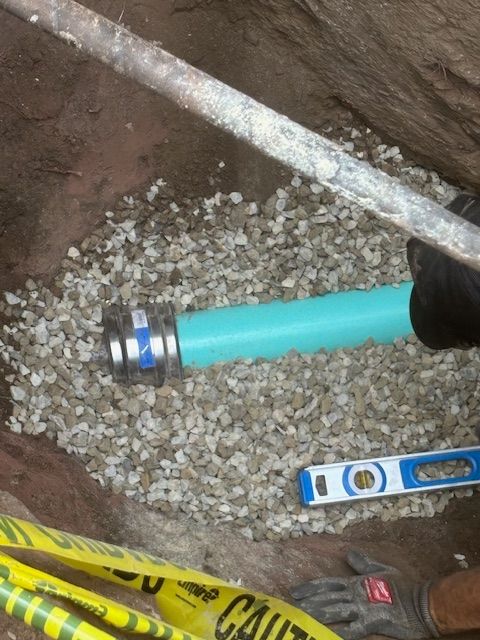 A turquoise pipe connection surrounded by gravel in an excavation, with a level and caution tape.