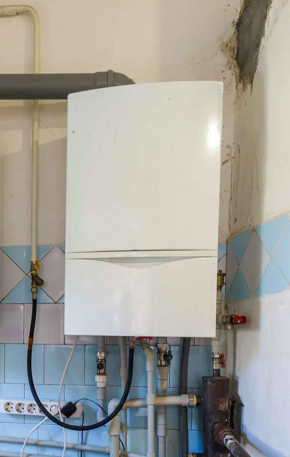 White wall-mounted gas boiler with pipes and electrical cord against blue and white tiled wall.