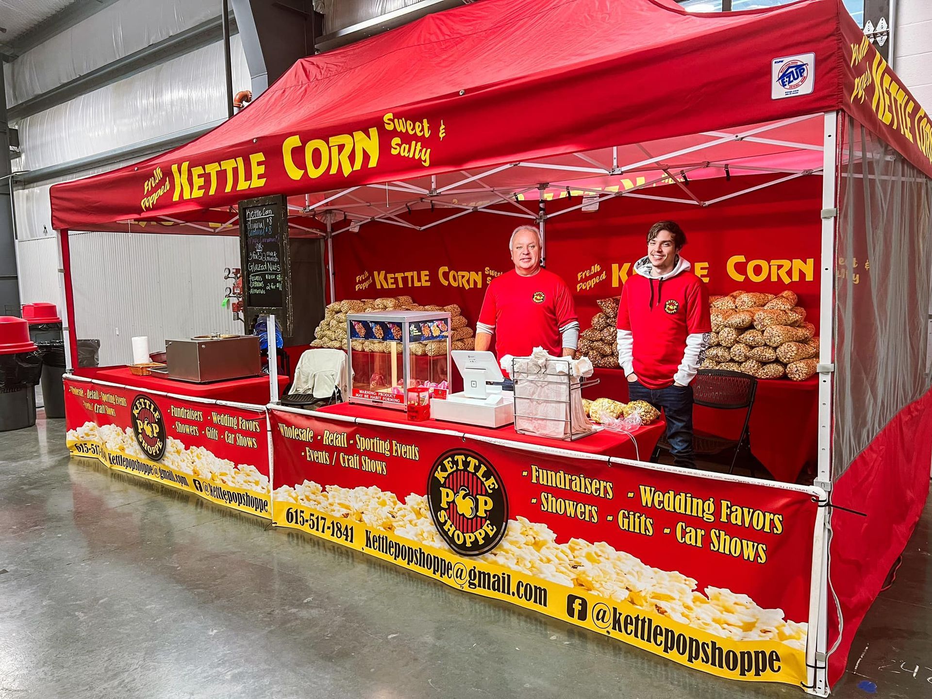 Two men are standing behind a popcorn stand that says kettle corn