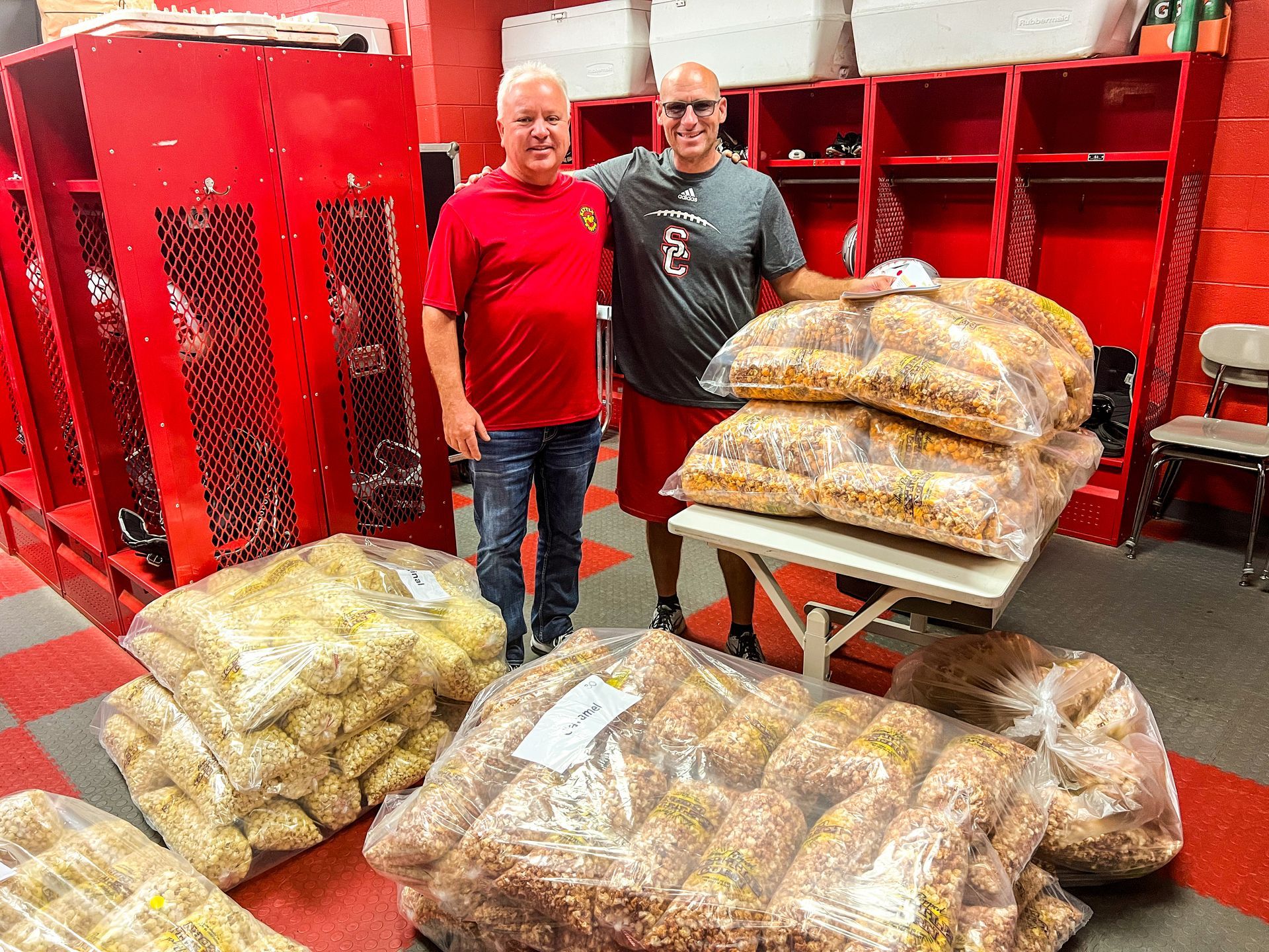 Two men are standing next to each other in a locker room holding bags of food.