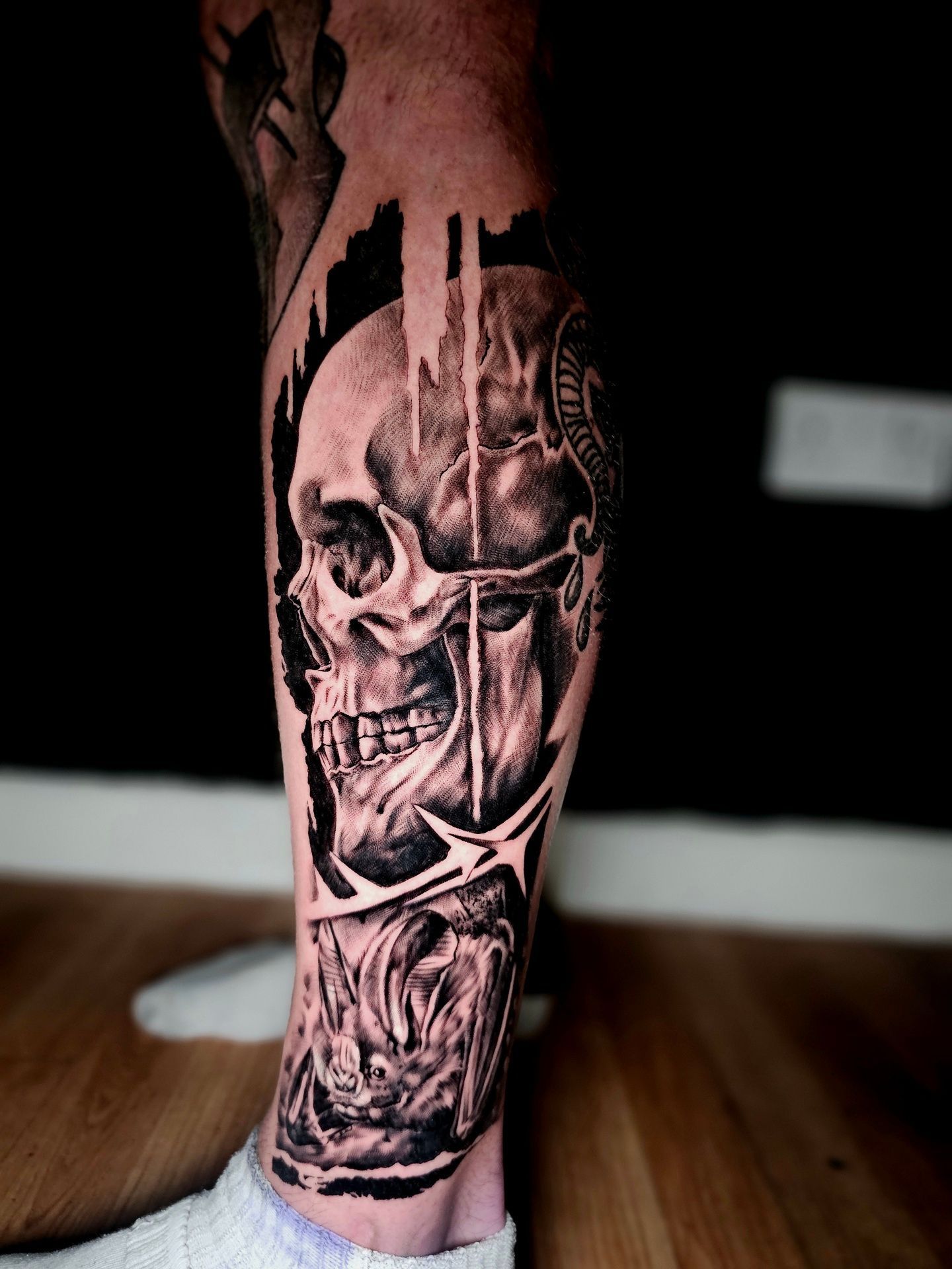 A black-and-grey tattoo on a lower leg featuring a detailed skull, abstract vertical lines, and barbed wire elements.