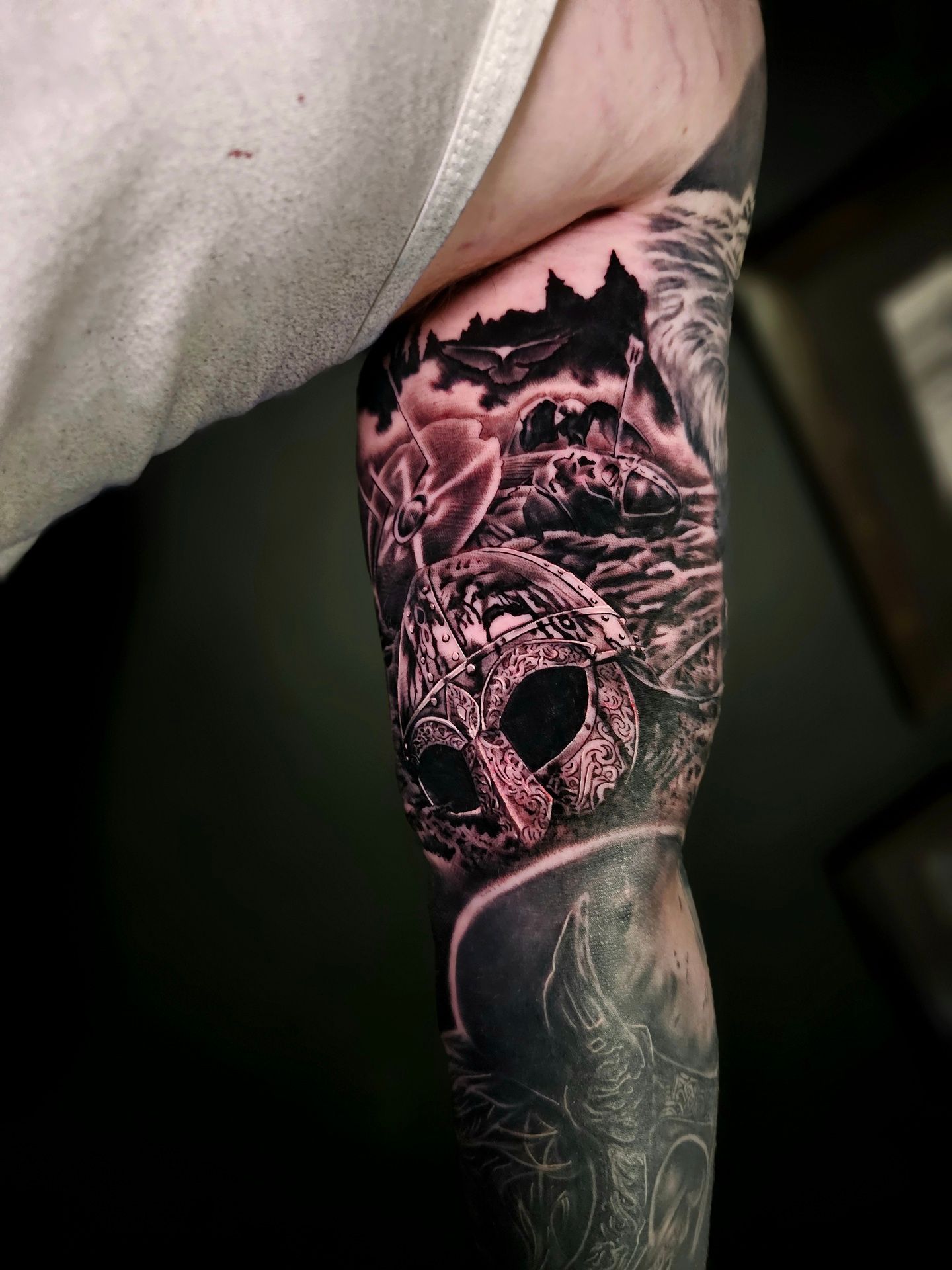 A detailed, black-and-grey tattoo on an inner arm featuring a spartan helmet, rocky mountains, and stylized background art.
