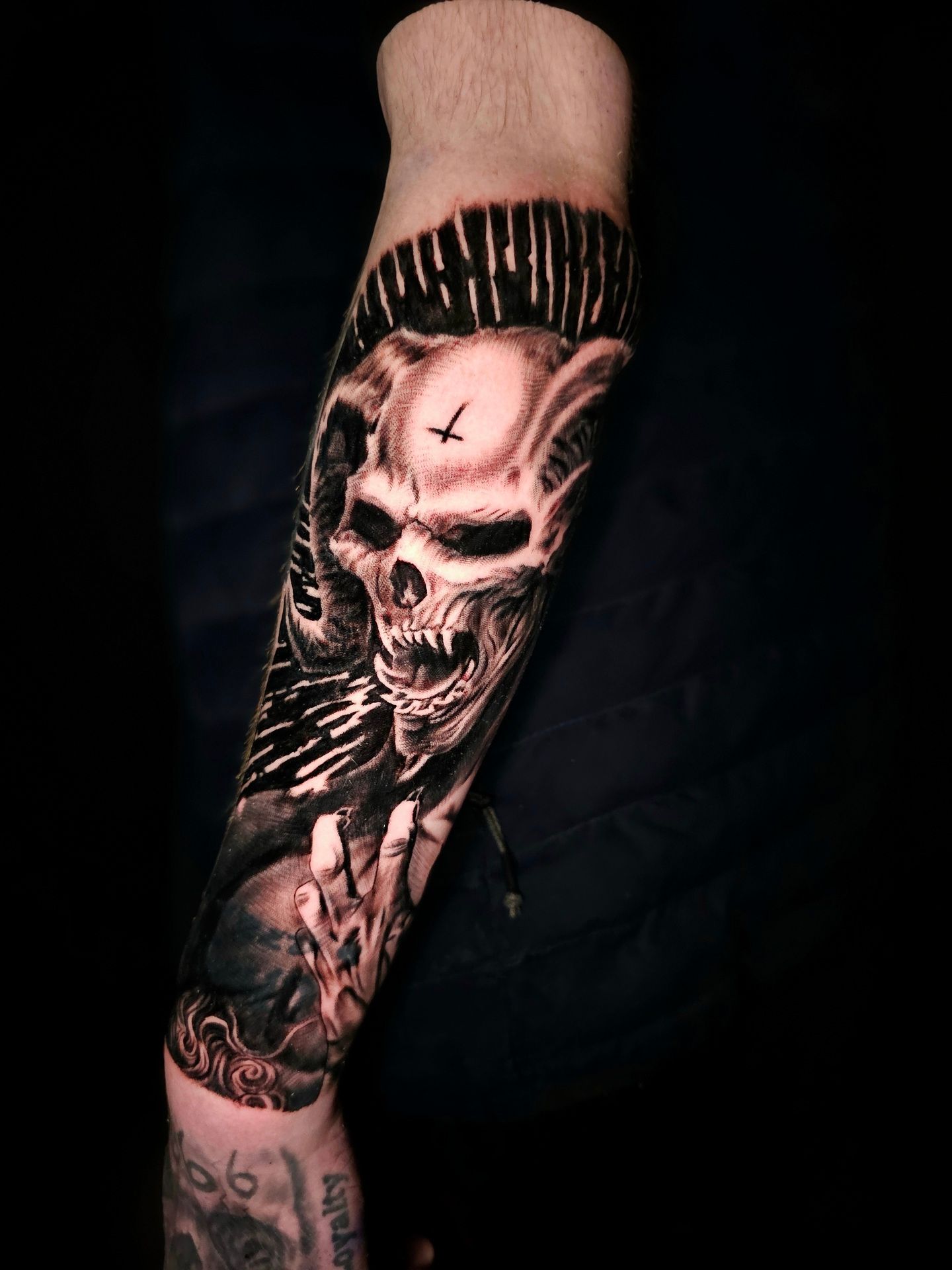 A forearm tattoo of a horned skull with a small inverted cross on its forehead, depicted in black and gray realism.