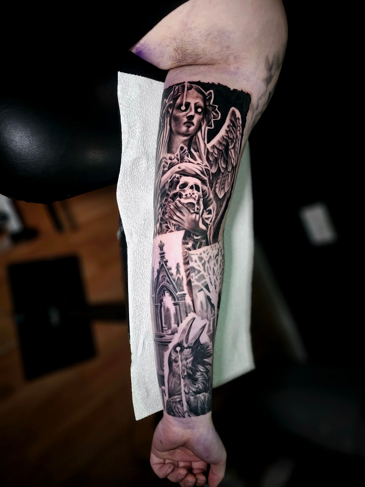A black-and-grey tattoo on a forearm featuring a gothic angel, a skull, and architectural elements like arches.