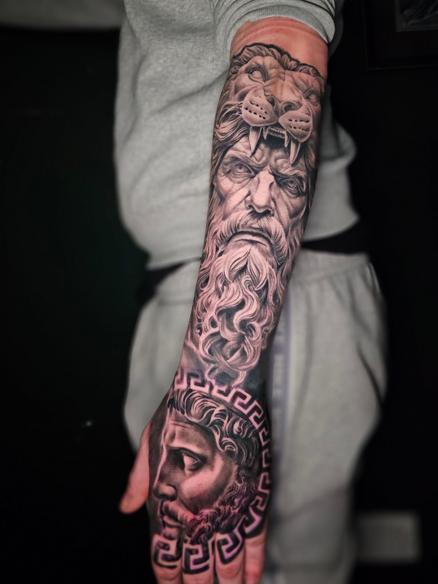 A forearm and hand featuring a detailed black and grey tattoo of a bearded man wearing a lion headdress and Greek motifs.