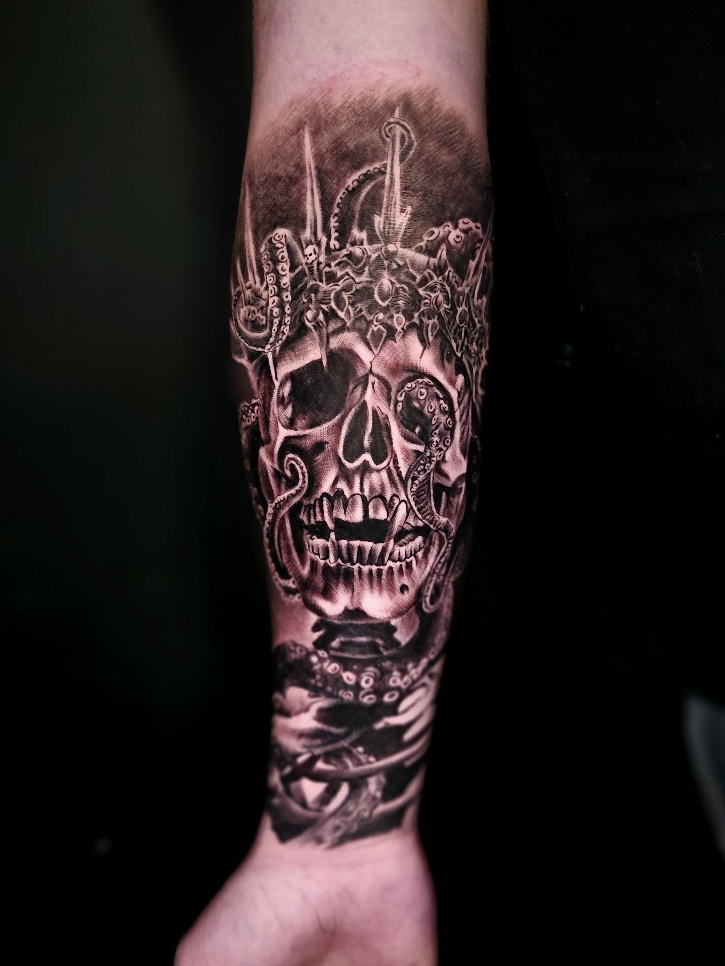 A black and gray tattoo on a forearm featuring a skull wearing a crown, surrounded by octopus tentacles.