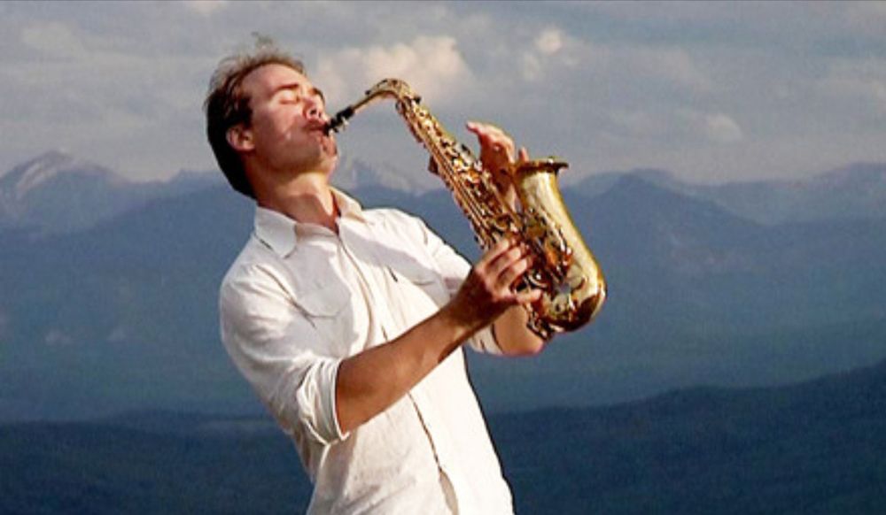 A man in a white shirt is playing a saxophone in front of mountains.