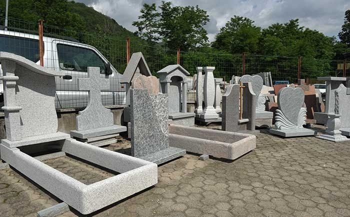 Un insieme di lapidi e segnacoli tombali in granito grigio disposti all'aperto su un piazzale pavimentato, con un veicolo sullo sfondo.