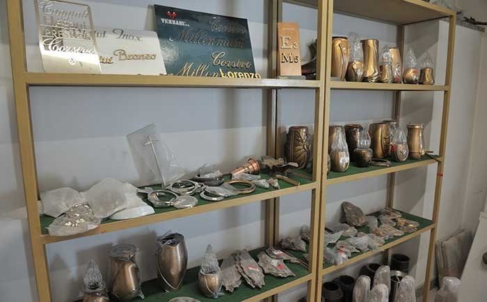 In un negozio, una scaffalatura metallica espone diverse urne funerarie, targhe commemorative e accessori decorativi per tombe.
