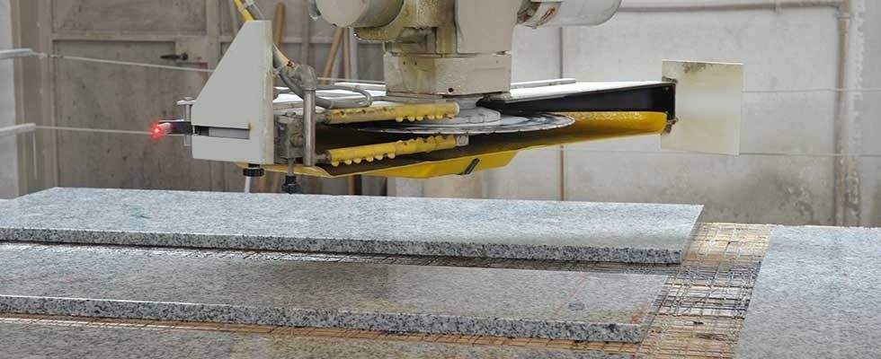 In un'officina di fabbrica, un braccio robotico con una lama circolare taglia una grande lastra di pietra grigia.